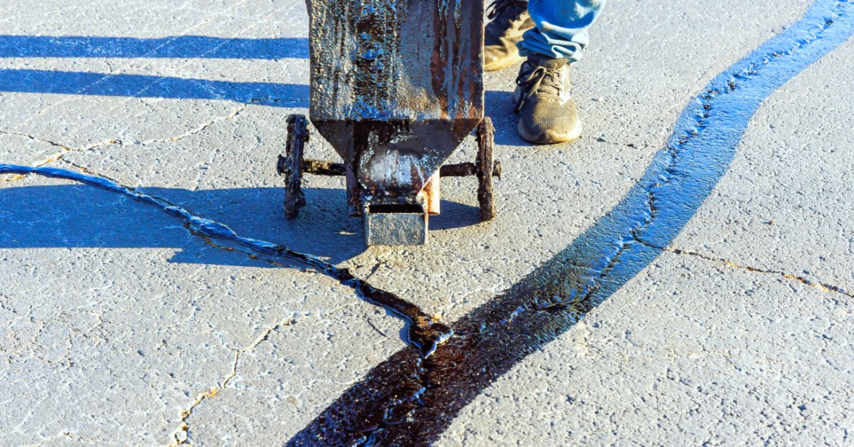 A wheeled asphalt sealing tool filling pavement cracks with dark material beside a person’s boots on a road.