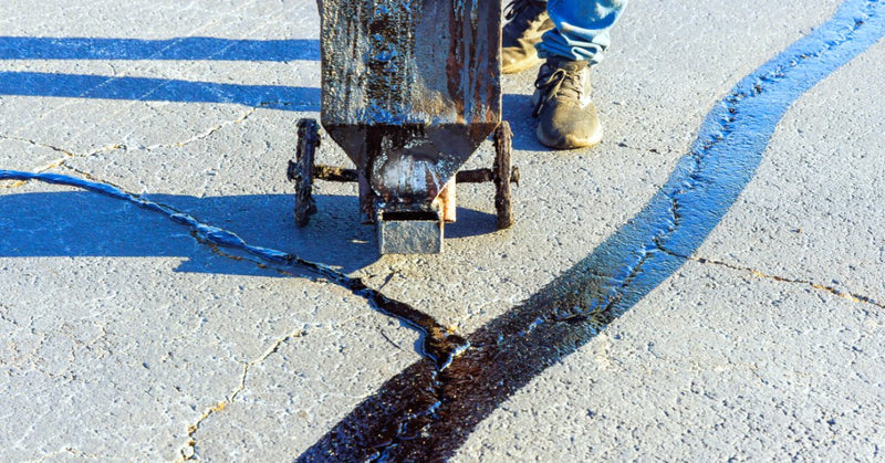 A wheeled asphalt sealing tool filling pavement cracks with dark material beside a person’s boots on a road.