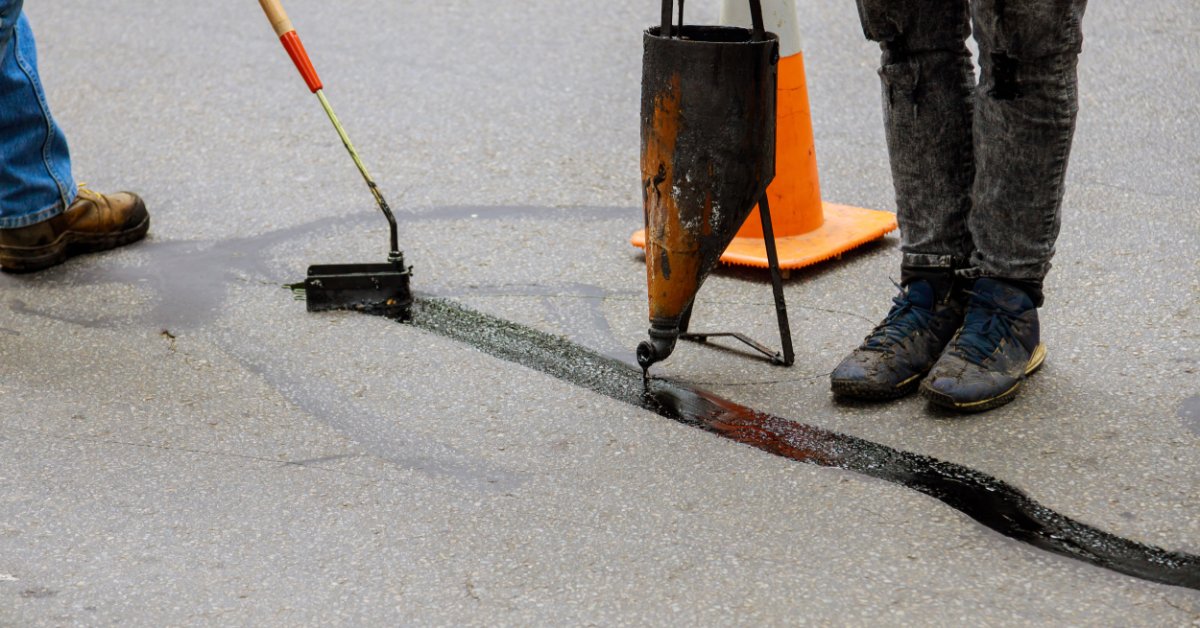 Workers using tools to pour and spread black sealant along a cracked asphalt road beside a nearby orange traffic cone.