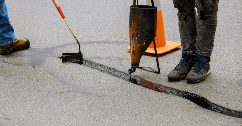 Workers using tools to pour and spread black sealant along a cracked asphalt road beside a nearby orange traffic cone.