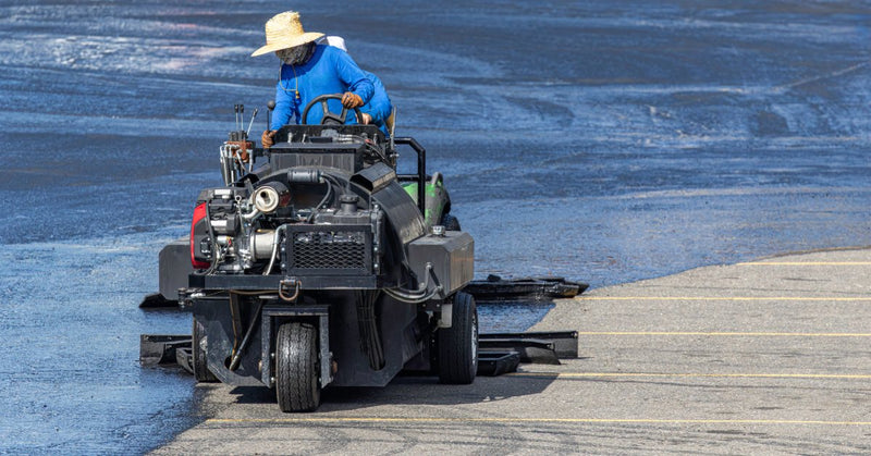 A worker wearing a straw hat operating a sealcoating machine across a parking lot with fresh blacktop.