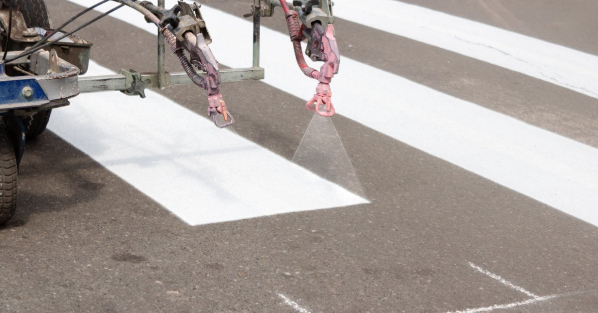 A striping machine applying thick white pedestrian crossing lines across dark asphalt with faint chalk markings.