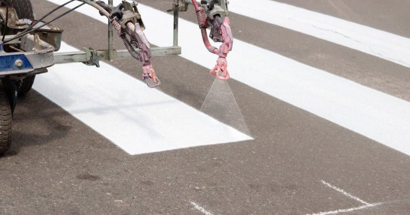 A striping machine applying thick white pedestrian crossing lines across dark asphalt with faint chalk markings.