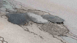 A close-up of a pothole in an old, cracked road. Some of the patches look older than others and have been re-patched.