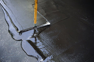 Someone sealcoating an asphalt surface using wet black sealcoating materials and a yellow sealcoating tool.