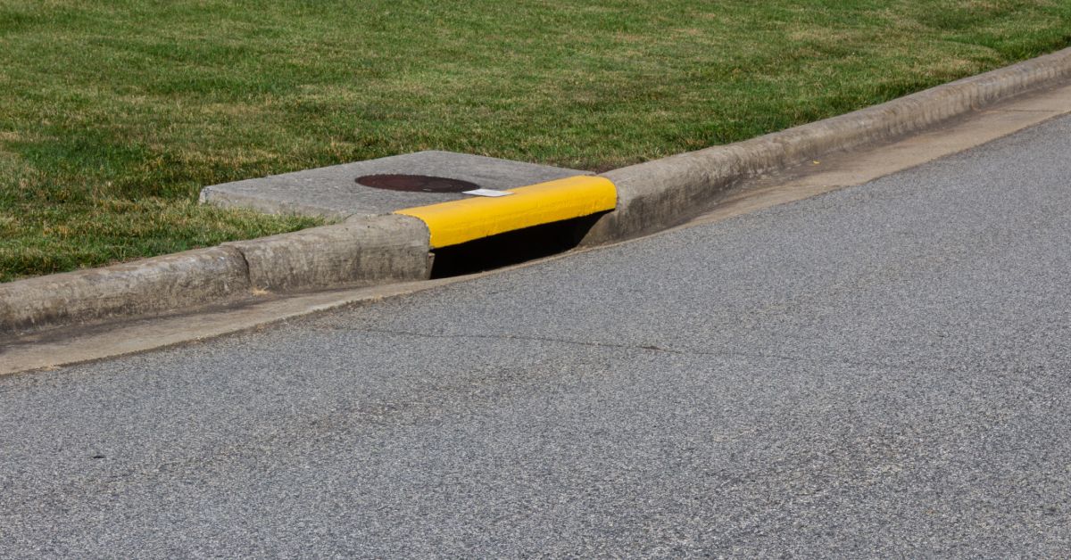 A residential street curb with an asphalt drain painted yellow next to green grass and an asphalt road.