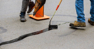 A close-up of two people's shoes as they do road restoration work. They're filling and sealing cracks with fresh asphalt.