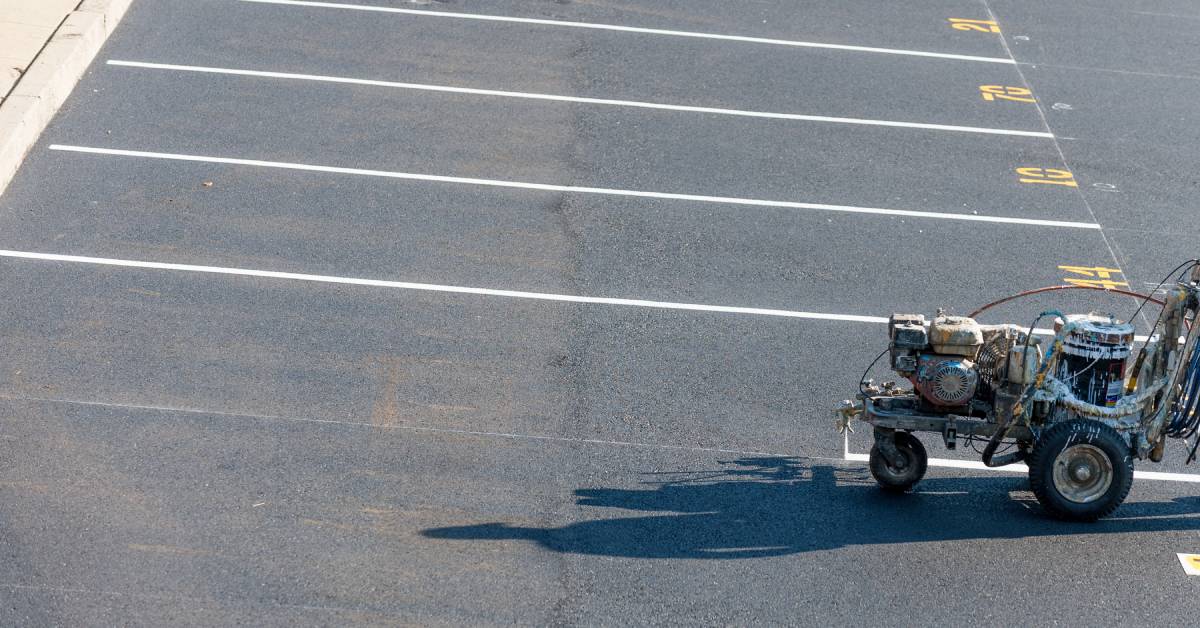 A silver striping machine painting clean white lines on an asphalt surface. The parking spots include yellow numbers.