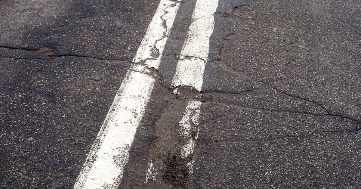 A cracked asphalt road with two solid white centerlines, showing wear and deterioration along the painted stripes.