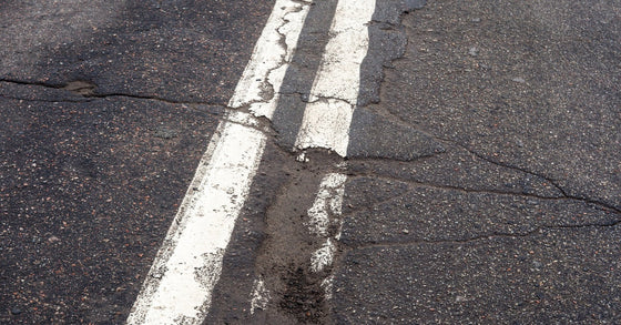 A cracked asphalt road with two solid white centerlines, showing wear and deterioration along the painted stripes.