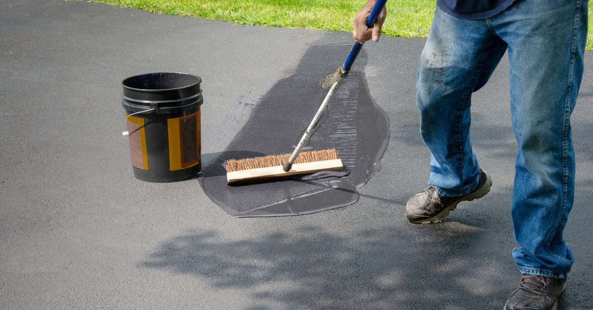 A person applying a thick gray sealant to a driveway using a squeegee/brush combo tool, with the bucket of sealant close by.
