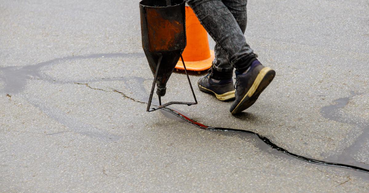 A person wearing jeans and sneakers using a crack filling machine to apply sealant to a fissure in the middle of a road.