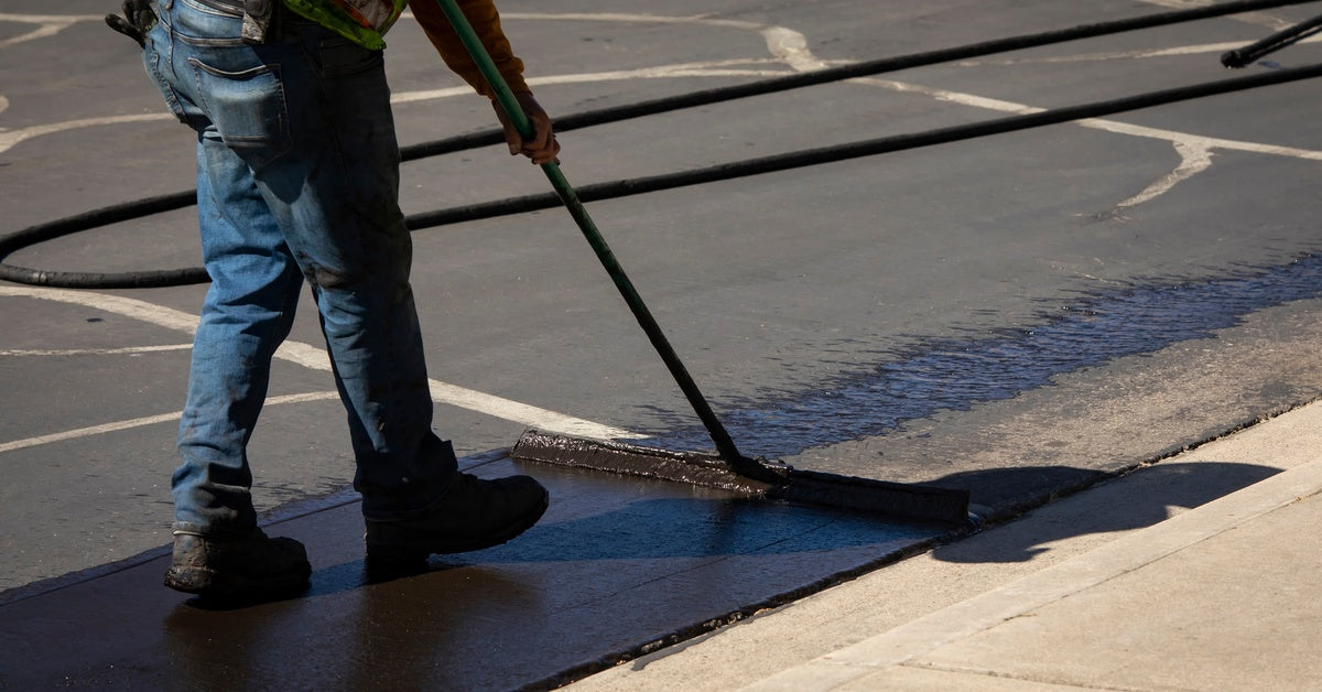 A person in a hi-vis vet, jeans, and work boots using a squeegee to spread sealcoat across an asphalt parking lot.