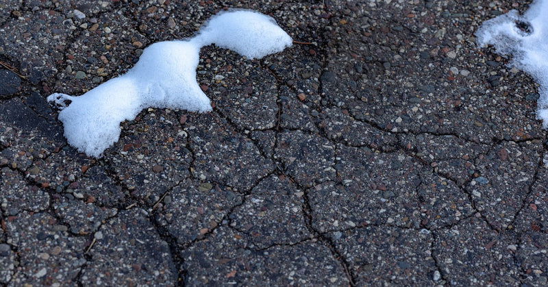 Asphalt pavement covered with dozens of small, interconnected cracks, along with small piles of snow on the surface.
