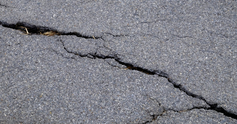 A close-up on worn-down asphalt pavement with multiple cracks and uneven texture visible throughout the surface.