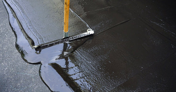 A long-handled squeegee spreading black sealant across a cracked asphalt driveway surface with visible streaks.