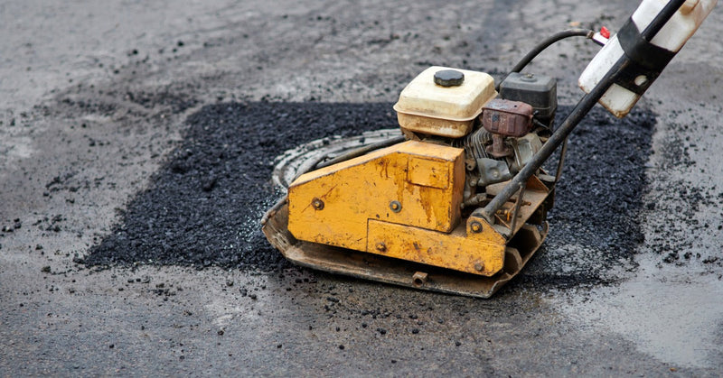 A yellow plate compactor pressing fresh black asphalt around a round manhole cover on a wet paved surface.