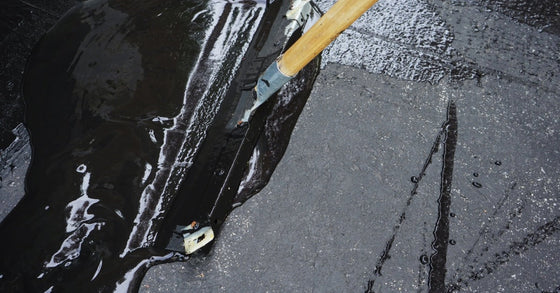 A long-handled squeegee spreading glossy black sealant across a gray asphalt driveway with visible cracks.