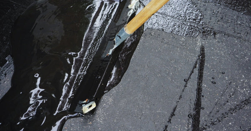 A long-handled squeegee spreading glossy black sealant across a gray asphalt driveway with visible cracks.