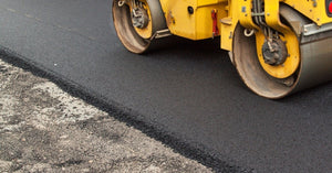 A yellow construction vehicle rolls over fresh black asphalt next to a section of road that is still gray.