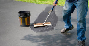 A man in blue jeans using the back of a broom to spread black sealant across an asphalt pavement with a bucket on it.