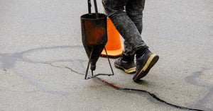 A man walks across asphalt pavement, dripping black crack sealing product from a piece of metal to repair a crack.