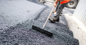 A worker in an orange safety vest pushes an asphalt lute front of him to level the application of fresh asphalt.