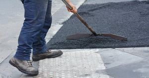 A person wearing blue jeans and brown boots rakes hot black asphalt beside a sidewalk with truncated domes.