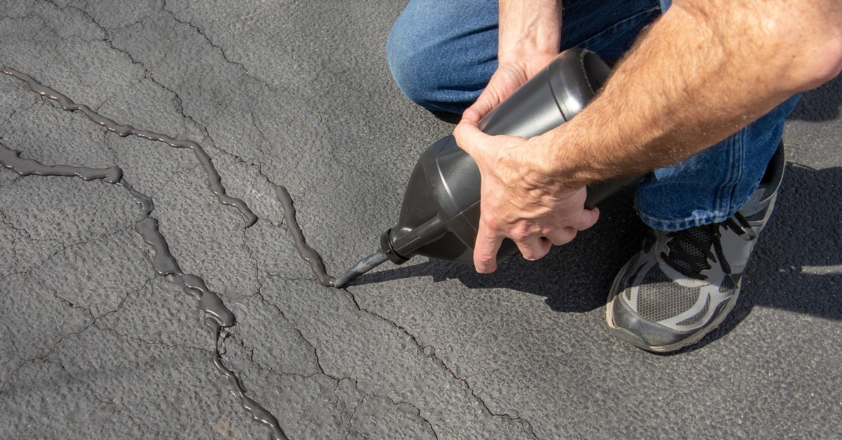 A man in blue jeans and tennis shoes repairs a crack in the asphalt by squeezing the liquid out of a black bottle.