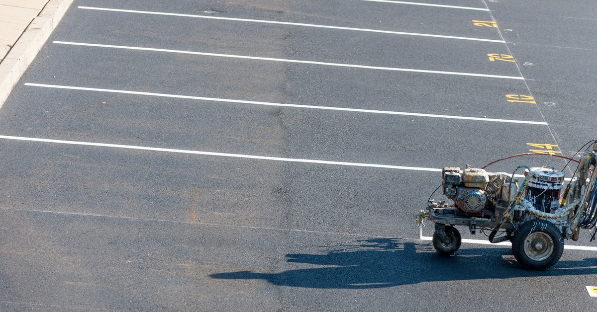 A parking lot striping machine is used over gray asphalt pavement to create white lines for parking spaces.