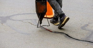A person wearing sneakers walks across asphalt pavement and seals a long crack with black liquid from a funnel.