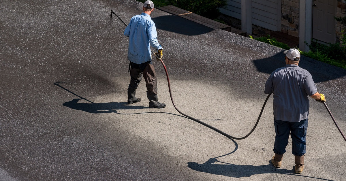 Two men in baseball caps hold a sprayer and spray as asphalt driveway with black sealant during a sunny day.