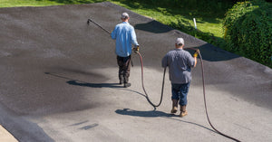 Two men walk across asphalt pavement with a spray gun hose, applying black asphalt sealcoat to the surface.