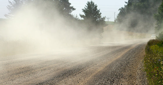 A gravel road with a lot of dust kicked up into the air. There is a car off in the distance at the end of the road.