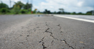 A grey asphalt street stretches out with many small cracks going through all of it into the distance under a blue sky.