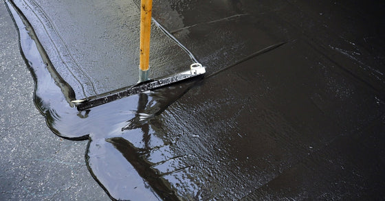 A close-up of a squeegee spreading black sealcoating liquid across an asphalt driveway to protect it from the elements.